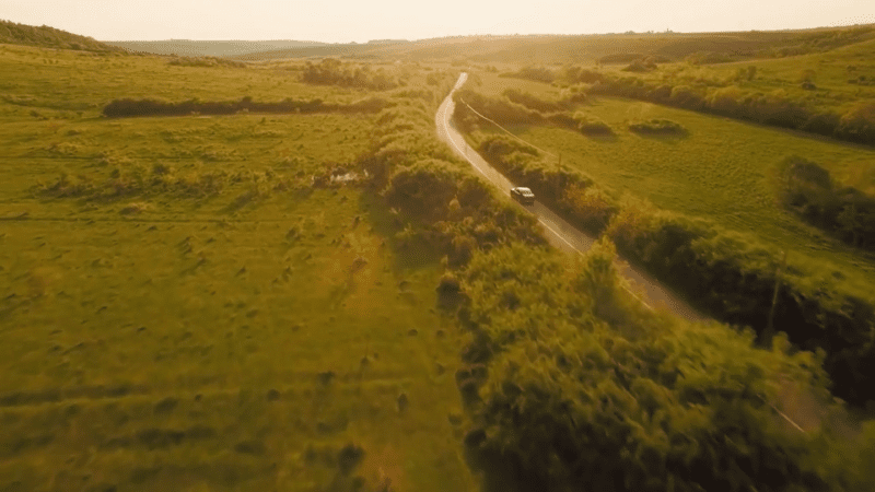 An aerial drone view of a lush, green landscape divided by a two-lane country road. A lone car can be seen driving between the hills under the late afternoon sun.